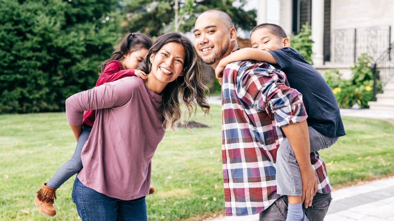 Family with two children, smiling, giving piggyback rides outdoors.