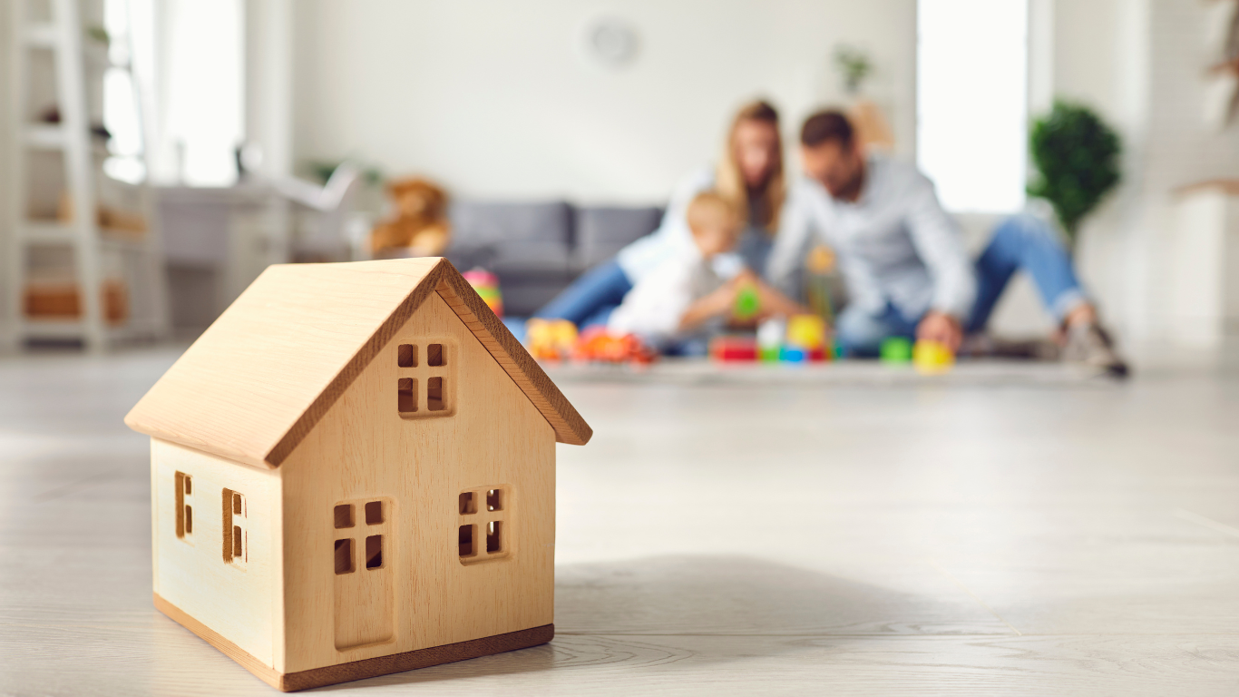 Wooden house model in focus with a blurred family playing in the background.