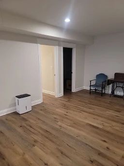 Basement with wood flooring, white walls, and a doorway. A dehumidifier and two chairs are in the space.
