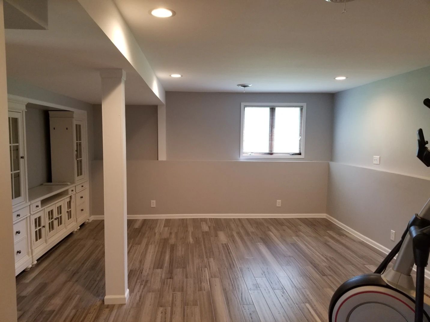 Empty basement room with gray walls, wood-look floor, window, and exercise equipment.