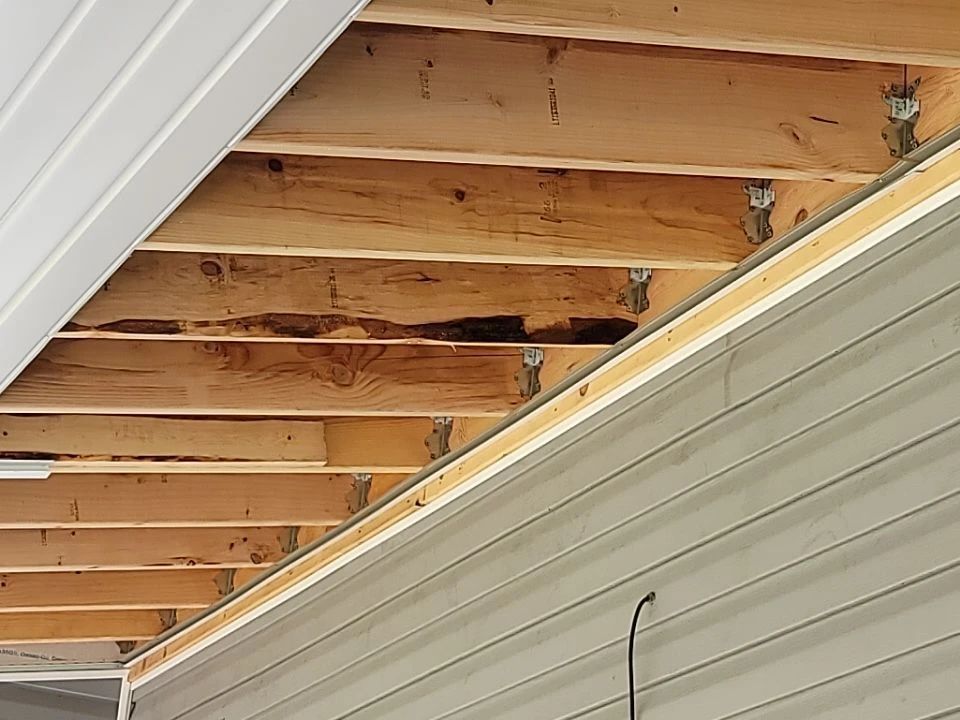 Underside of a wooden overhang with visible water damage, some boards missing. Gray siding and white trim.
