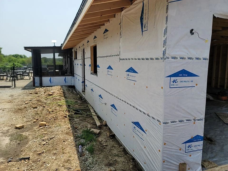 Exterior house under construction with white protective wrap and unfinished wooden framing.
