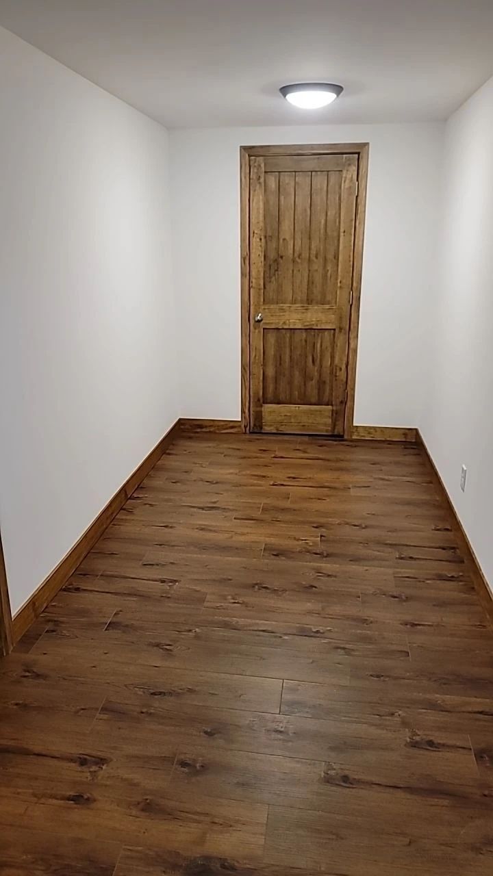 Narrow hallway with wood-look flooring and a wooden door at the end; white walls, and a ceiling light.