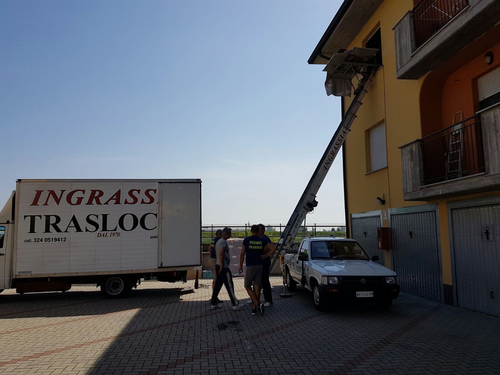 Un camion per traslochi scarica mobili dalla finestra di un edificio utilizzando un sollevatore. Tre persone sono in piedi vicino al camion e all'auto.
