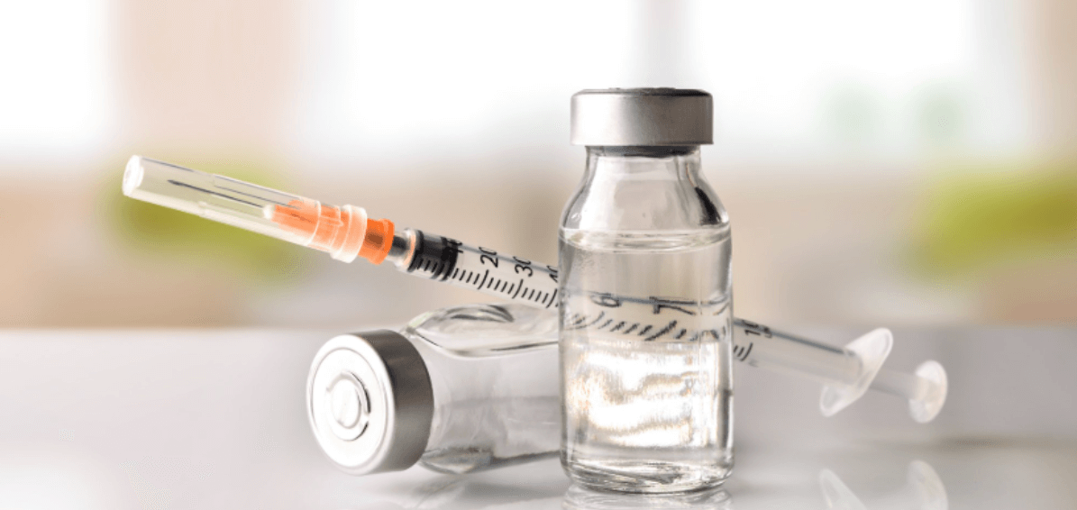 Syringe beside a glass vial of medicine on a bright tabletop, with a blurred background