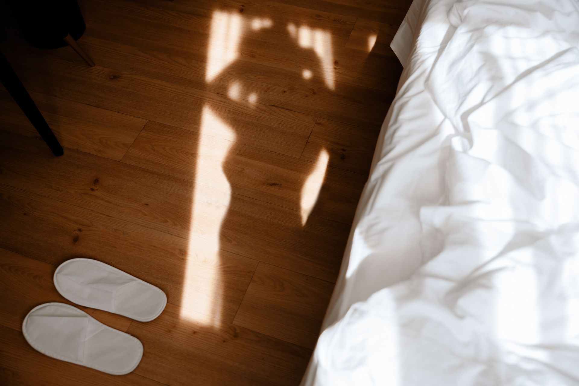 White slippers on a wooden floor beside a bed, with a bright shadow cast by sunlight.