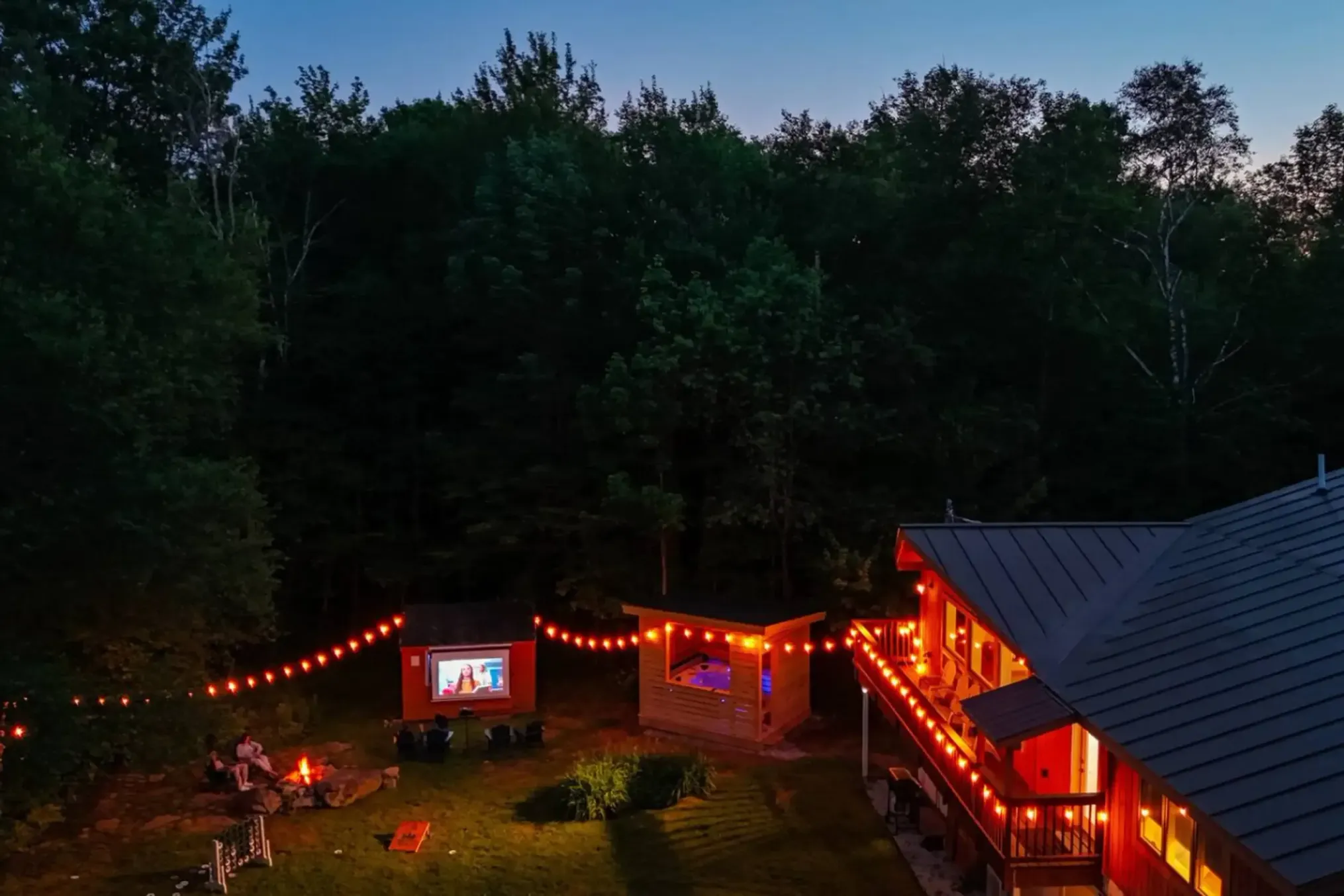 Backyard movie night by a fire pit at dusk with a lit screen, jacuzzi, string lights, and a house deck glowing red-orange