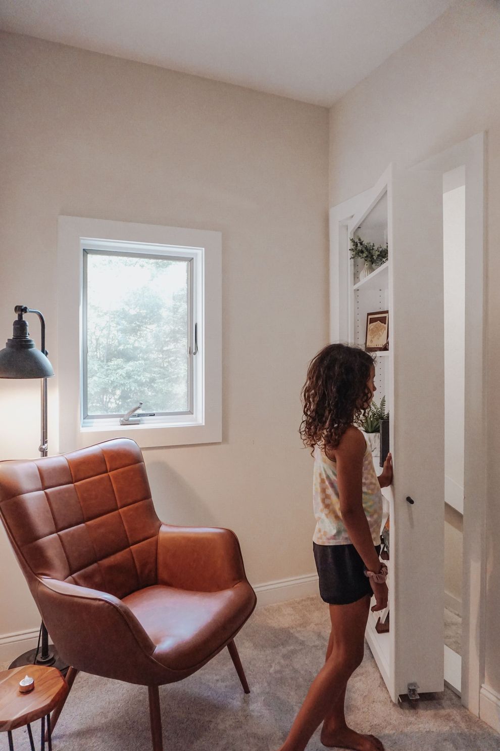 Person standing beside a bookcase that's a secret door in a bright room with a brown chair and window