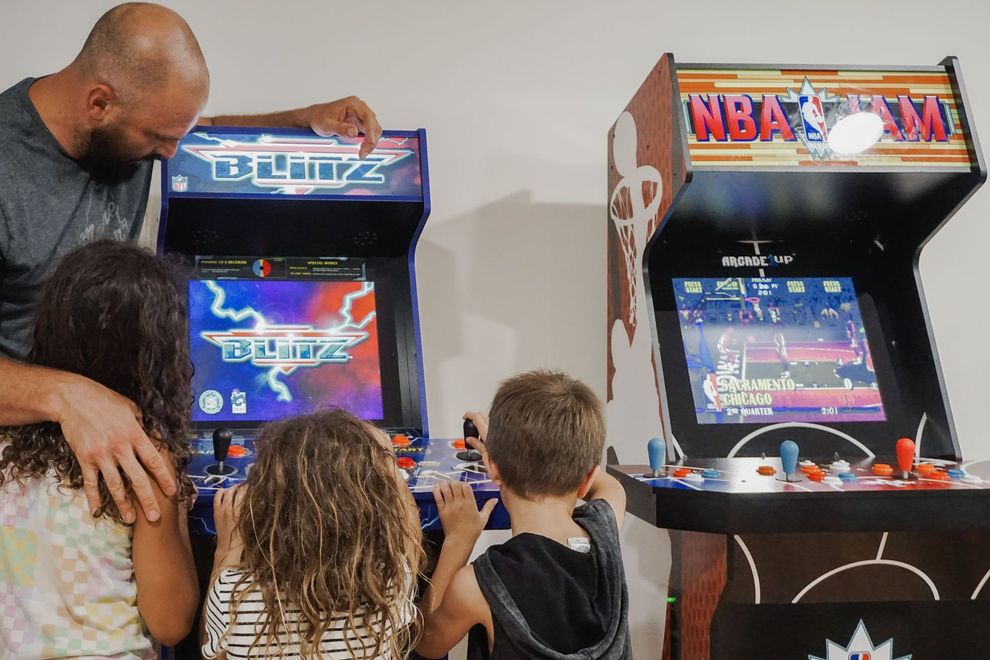 Two people and two children play arcade basketball games beside a black cabinet in a game room