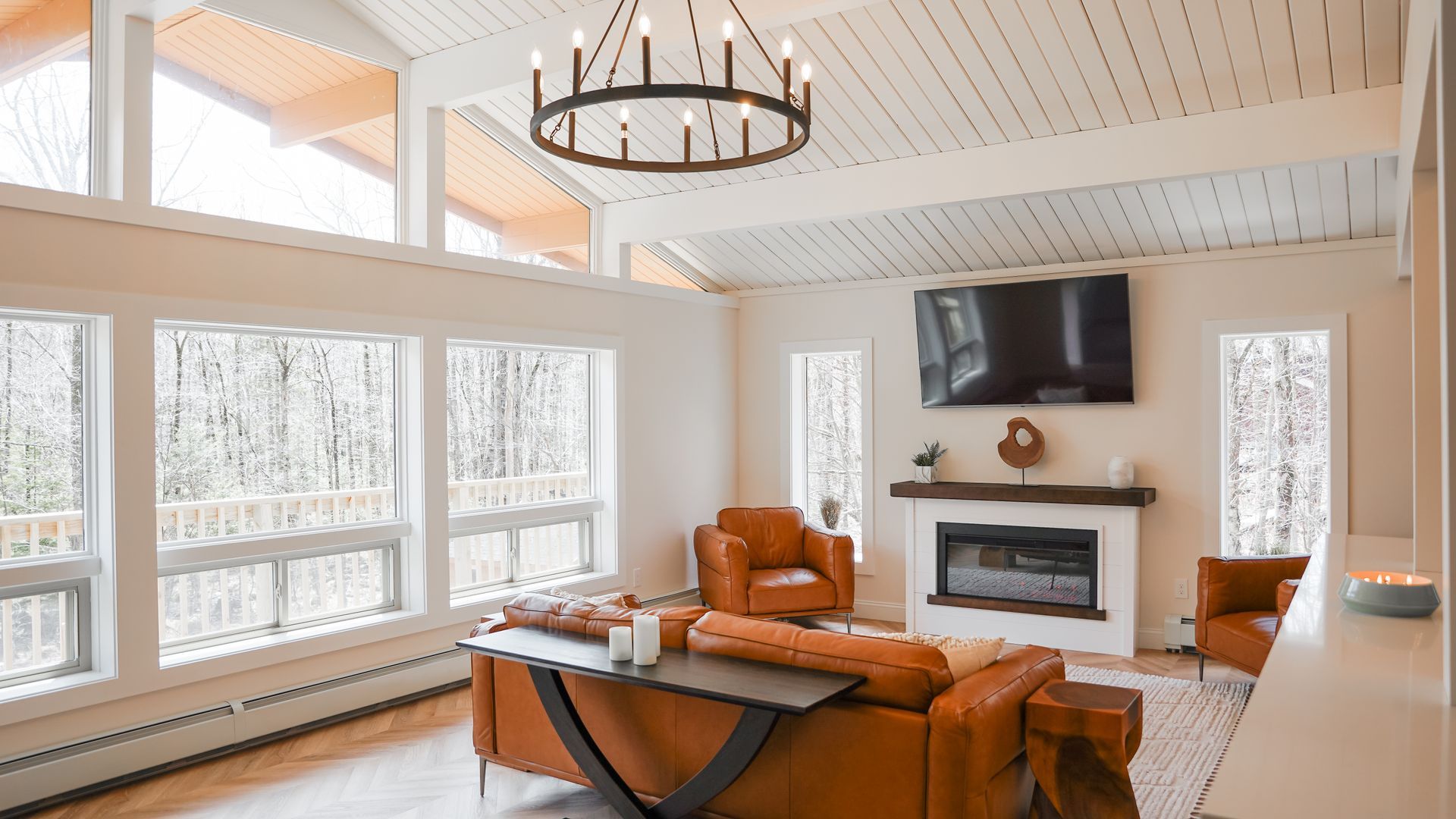 Bright living room with wood ceiling, leather sofas, a fireplace, wall-mounted TV, and large windows overlooking snowy trees