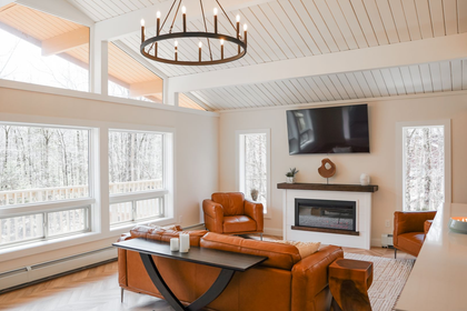 Bright living room with fireplace, wall TV, orange chairs, large windows, and a leather sofa.