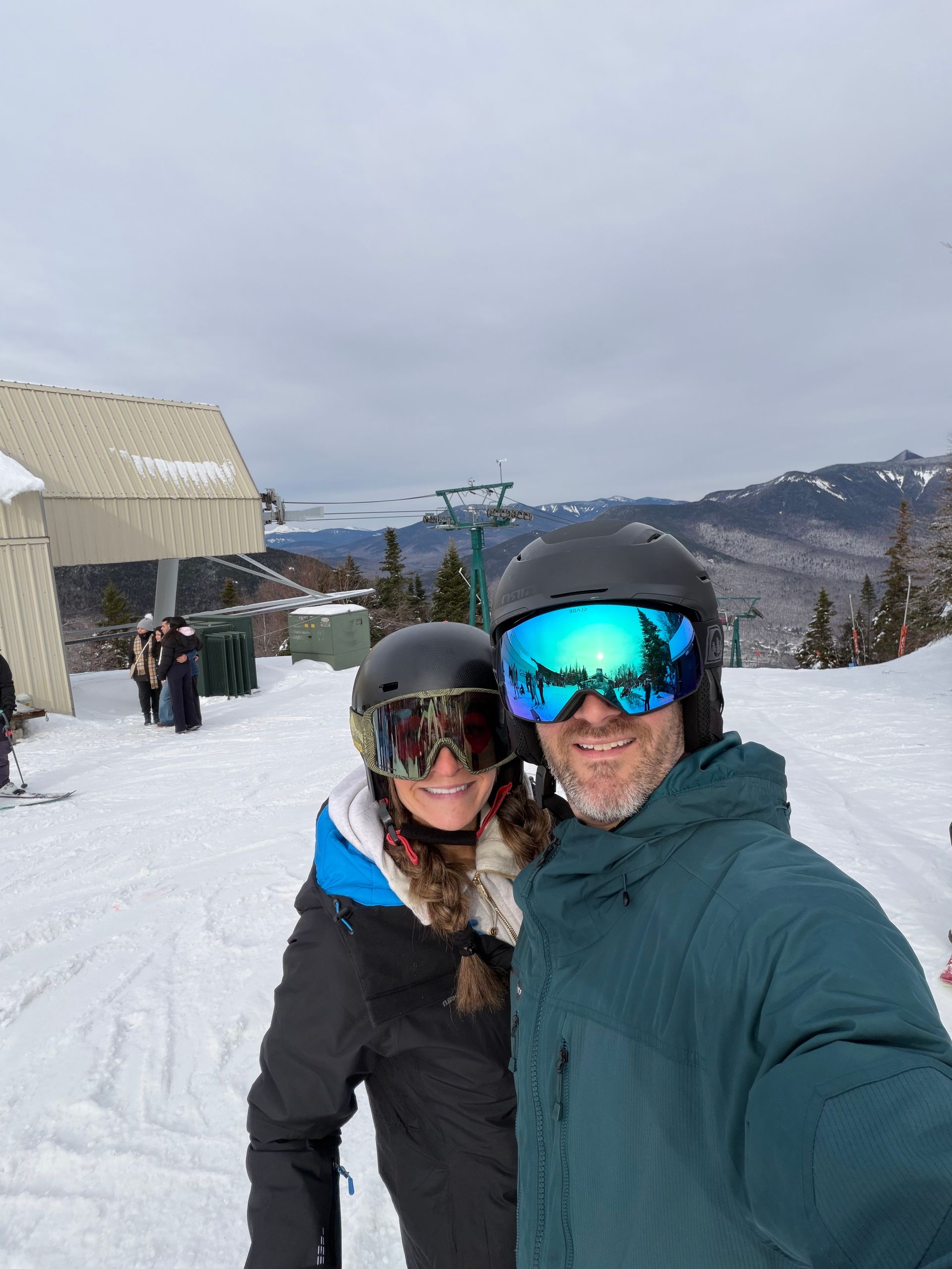 Two people in ski gear posing on a snowy mountain slope, with snowy hills and a cloudy sky behind them.