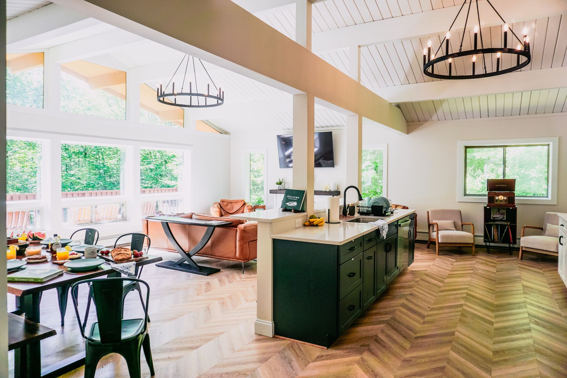 Bright open-plan living room and kitchen with white beams, large windows, wood chevron floors, and modern furnishings