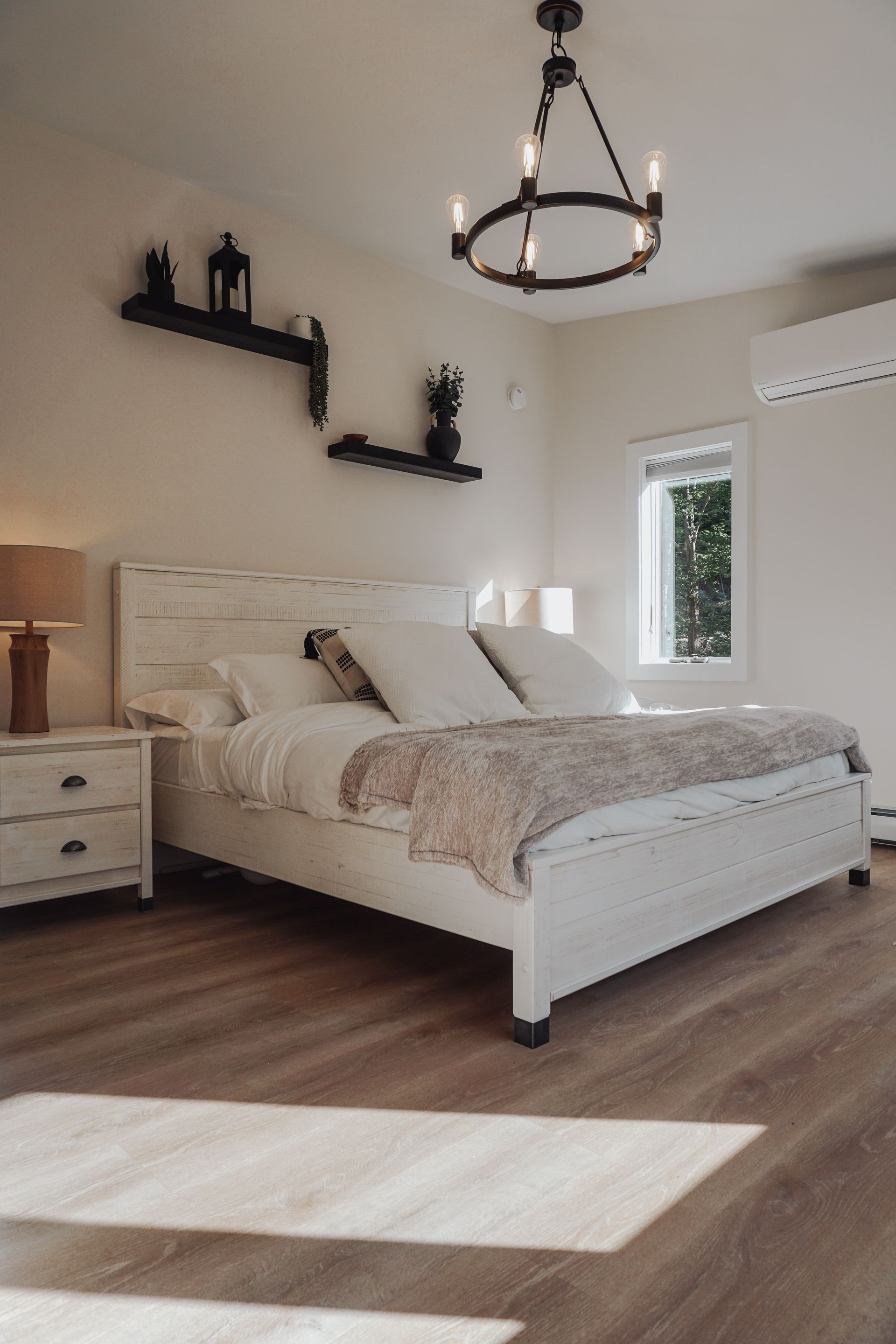 Bright bedroom with white bed, black wall shelves, pendant light, and sunlight on the floor.