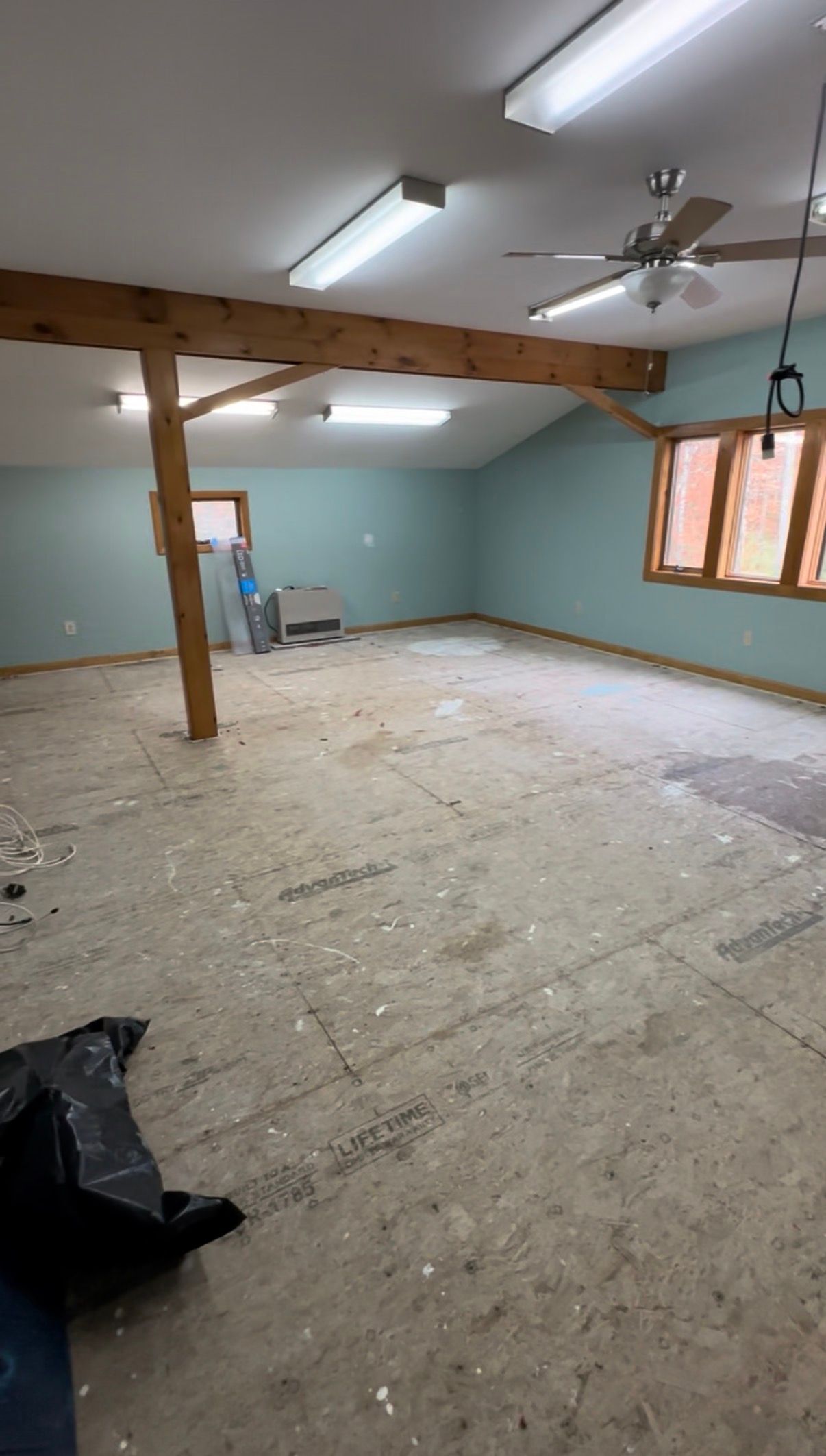 Unfinished empty room under renovation with teal walls, exposed floor, ceiling beams, and a window.