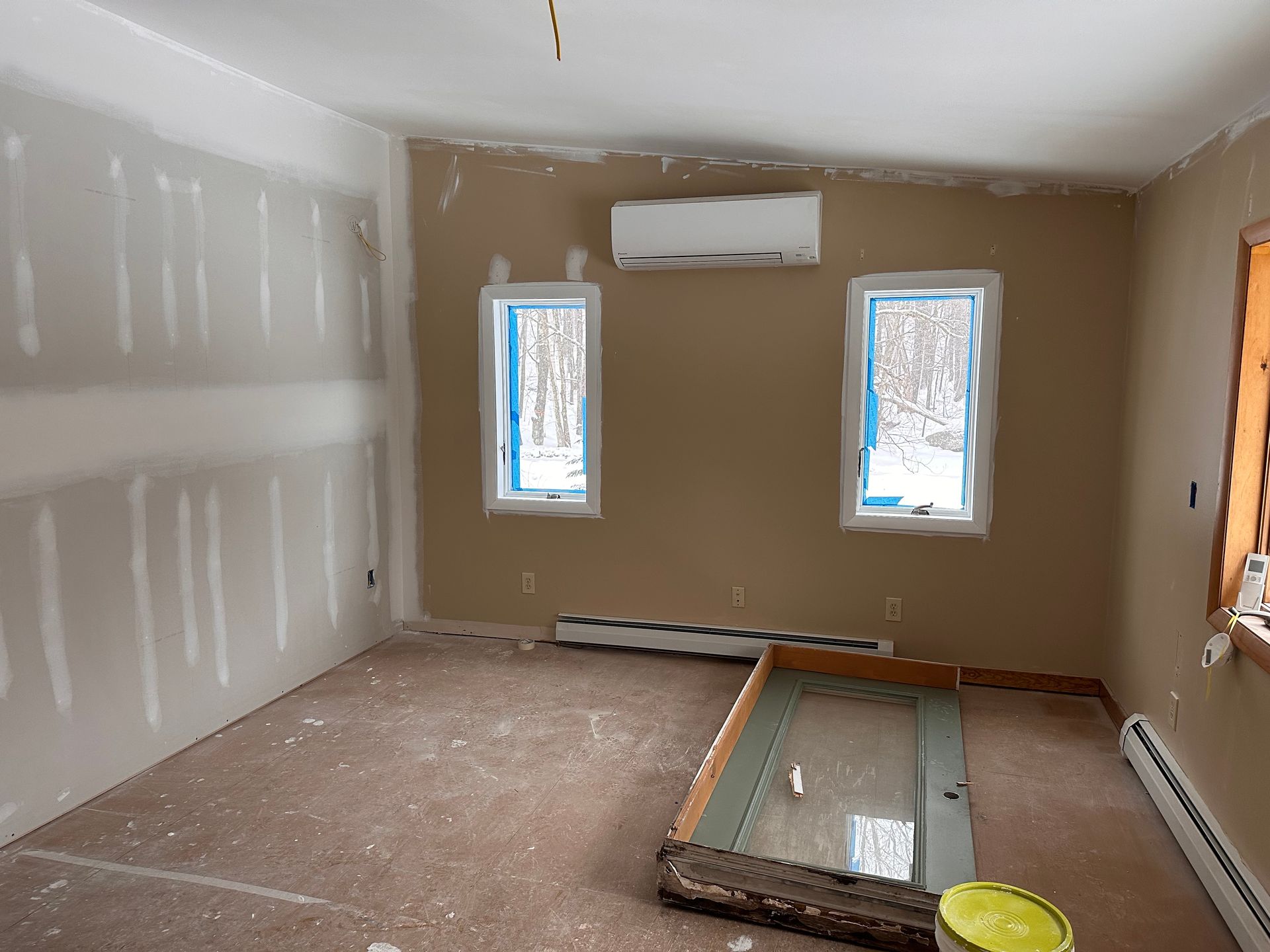 Empty room under renovation with drywall, two windows, a wall AC unit, and construction materials on the floor