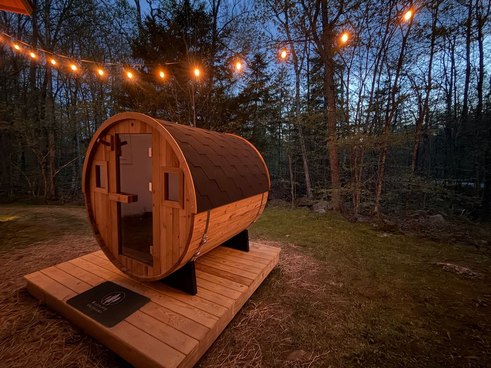 Wooden barrel-shaped sauna on a deck at dusk, lit by hanging string lights in a wooded yard