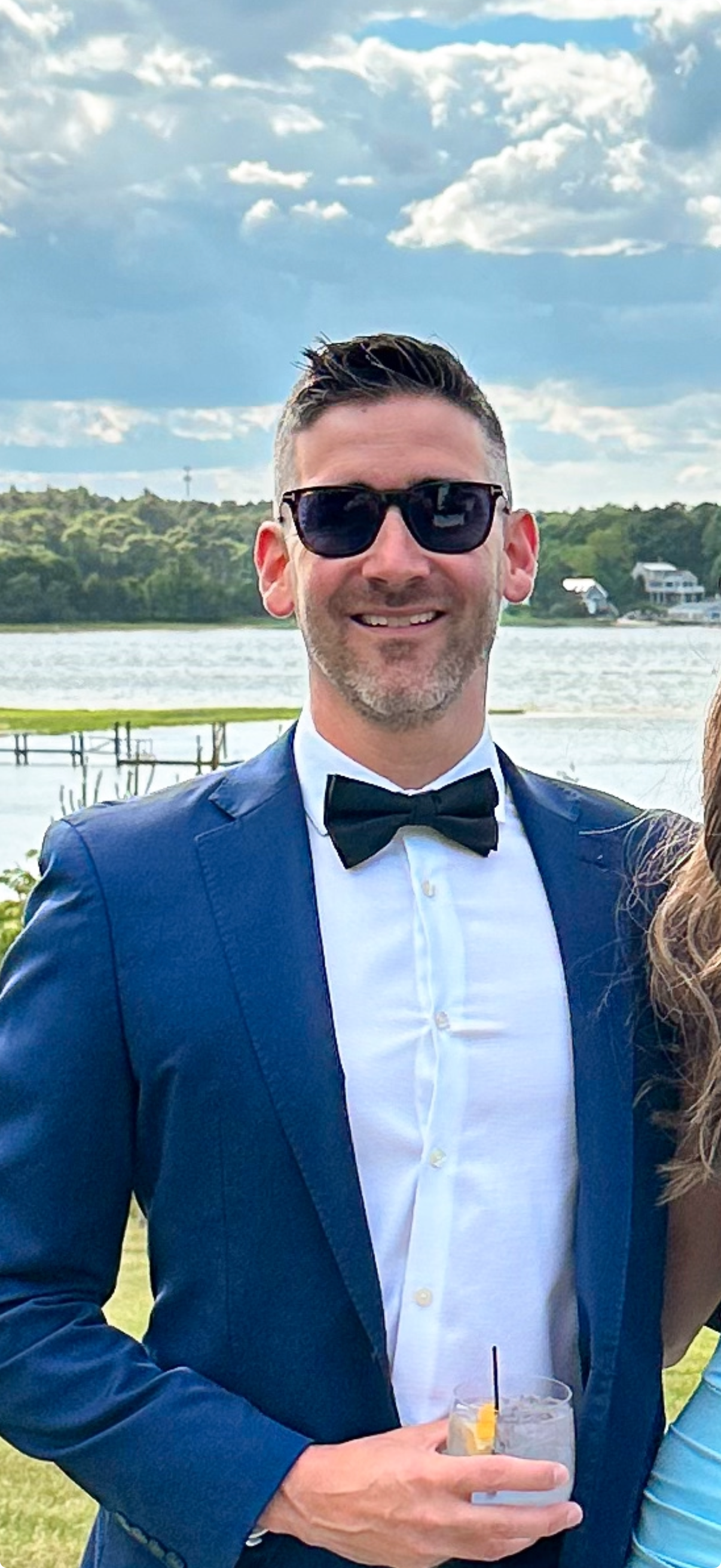 Man in blue suit and sunglasses smiling outdoors by the water at a formal event