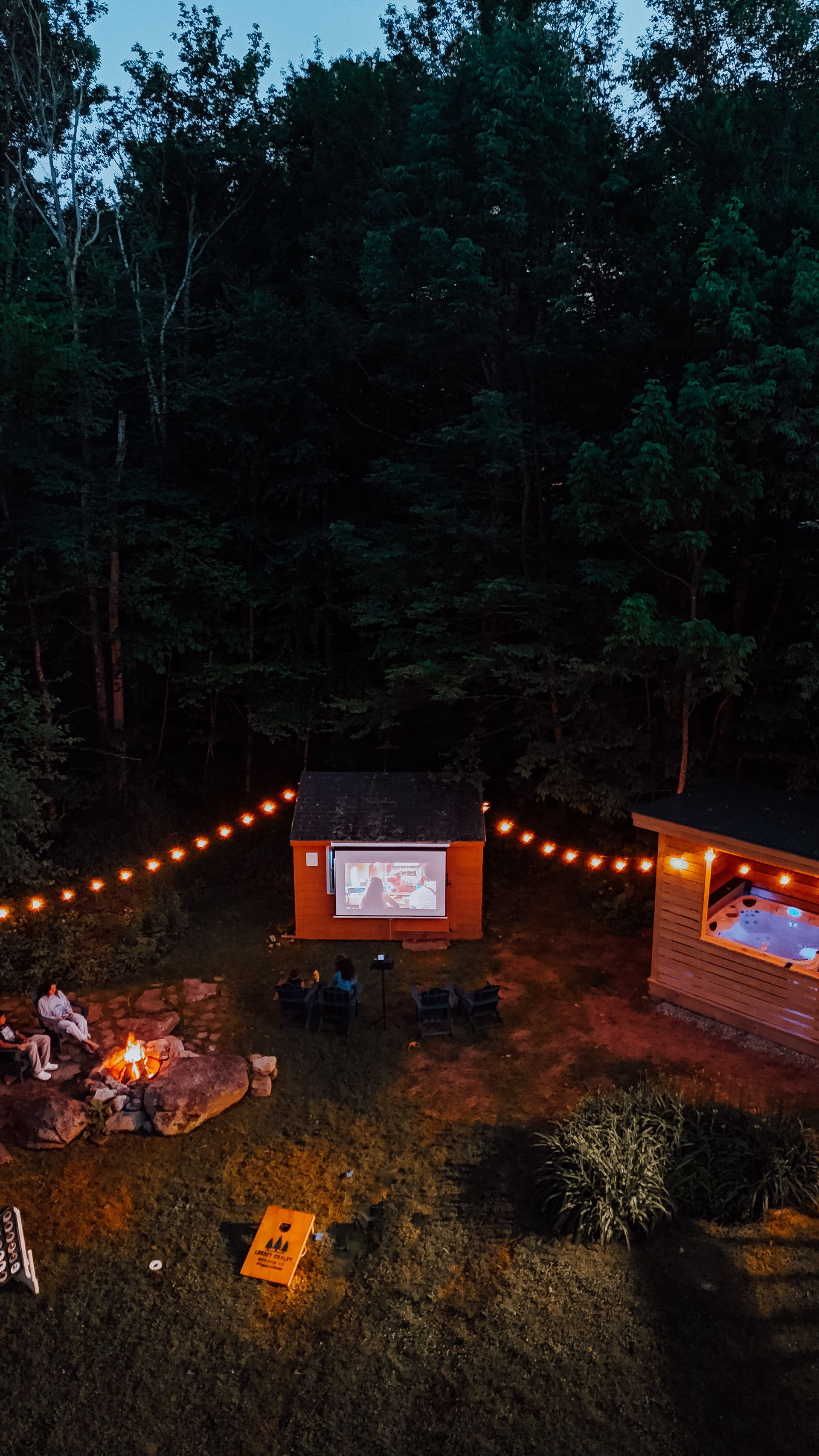 String-lit outdoor movie setup in a wooded clearing with a campfire and a projection screen.