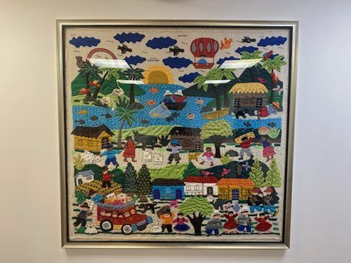 Colorful folk art village scene with houses, people, trees, clouds, and a red truck in a framed textile.