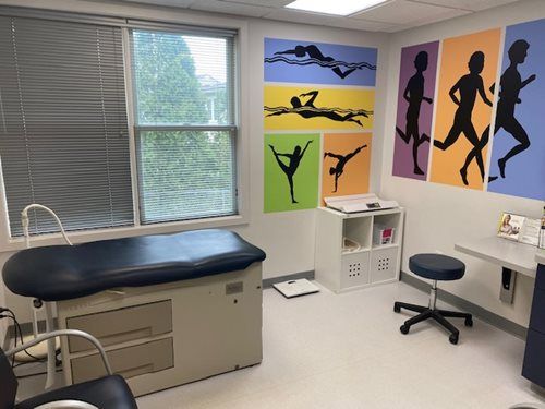 Medical exam room with exam table, stool, window blinds, and colorful sports silhouette wall art.