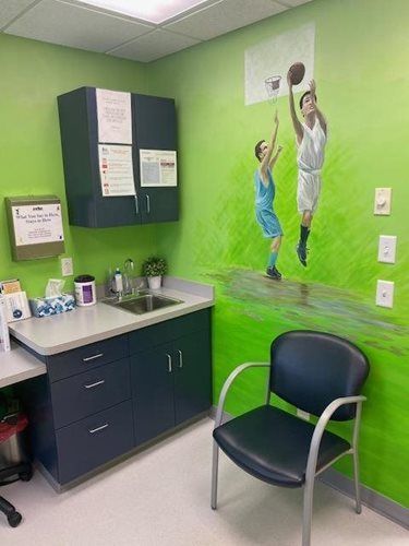 Small clinic room with sink, cabinets, chair, and a mural of children playing on a green wall.
