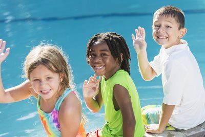 Three children smiling and waving by a bright blue pool on a sunny day