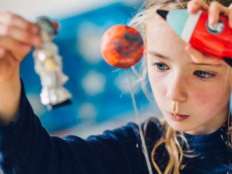 Child holding a small model of a red-and-white space scene, focused on a hanging planet craft project