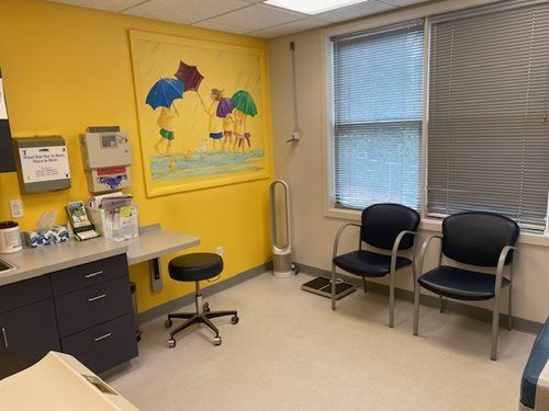 Small medical exam room with yellow wall, desk, stool, chairs, window blinds, and umbrella wall art