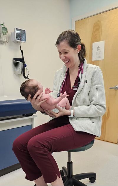 Healthcare worker holding a baby in a clinic exam room, both smiling on an exam chair.