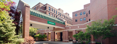 Brick hospital entrance with covered walkway and green sign, framed by trees and red brick buildings