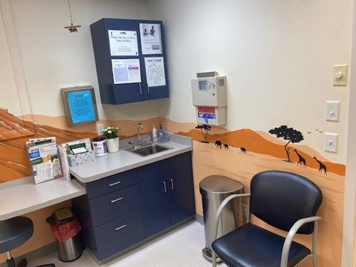 Small medical exam room with sink, cabinets, wall chart, hand sanitizer, and a chair by a counter.