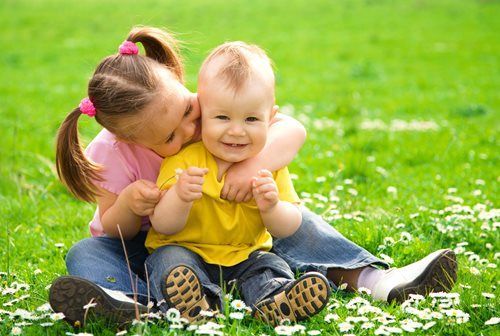 Two children sitting on grass, smiling and hugging in a sunny park