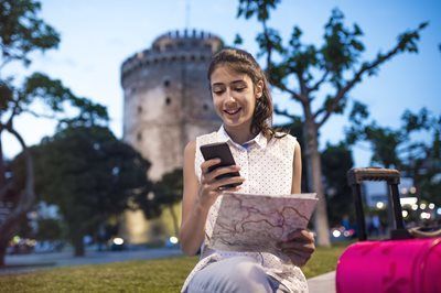 Person sitting on grass, smiling at a phone and map near a lit historic tower at dusk.
