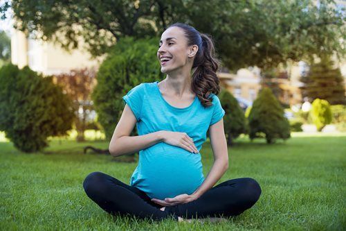 Pregnant woman sitting cross-legged on grass, smiling and holding her belly in a park
