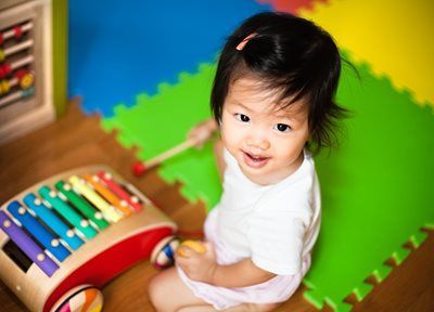 Toddler playing a colorful xylophone on a foam mat, smiling up at the camera.