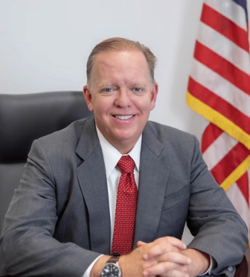 Man in gray suit and red tie seated at a desk with a U.S. flag in the background
