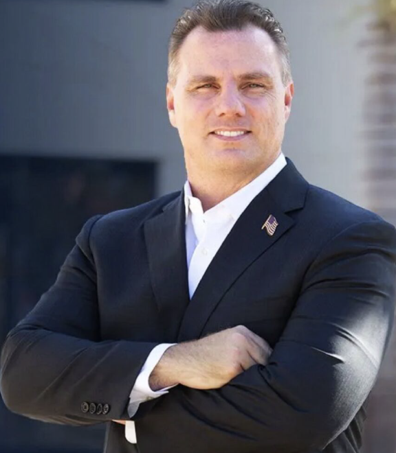 Person in a dark suit with arms crossed, smiling against a blurred outdoor background.