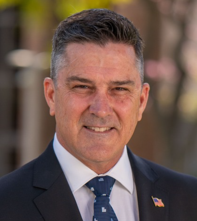 Portrait of a man in a dark suit and blue tie against a blurred outdoor background