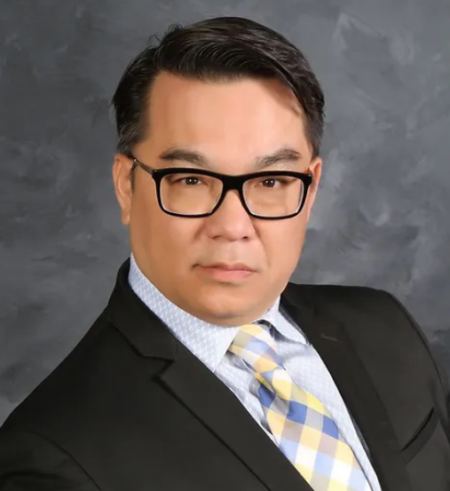 Studio headshot of a person in a black suit, striped shirt, and yellow tie against a gray background