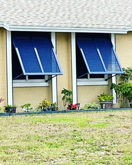 Two windows on a tan building exterior with open, blue metal storm shutters and various potted plants at the base.