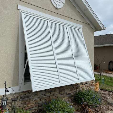 A white, three-paneled hurricane shutter is propped open over a window on a house with a tan stucco and stone exterior.