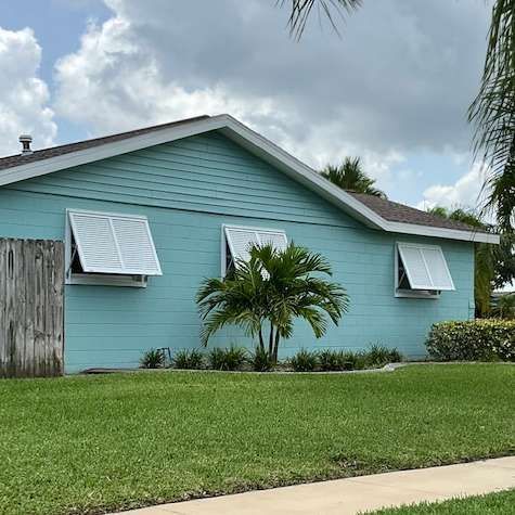 A light blue, single-story house with white awning-style windows, a brown roof, and a small palm tree in the front yard.