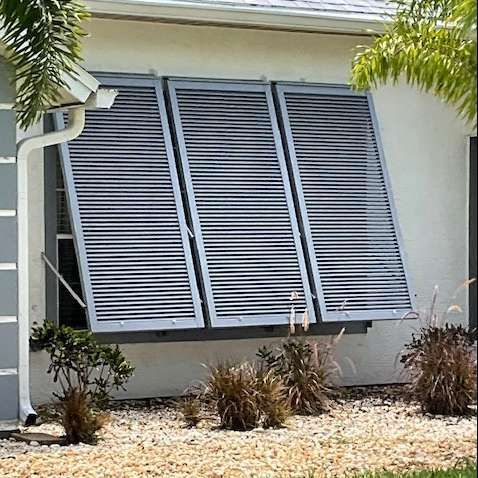 Three gray metal accordion-style storm shutters are deployed over a house window above a rock garden.