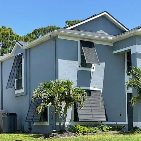 A two-story blue house with white trim and dark, angled metal hurricane shutters deployed over several windows.