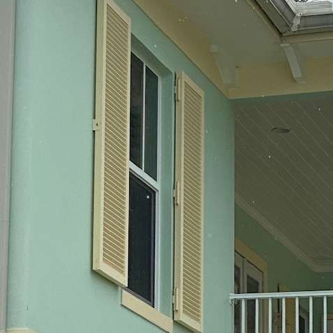 A window on a mint-green exterior wall featuring light yellow louvered shutters and a balcony railing visible nearby.