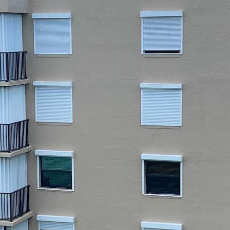 An exterior wall of a tan building featuring a vertical column of balconies and rows of windows with rolling shutters.