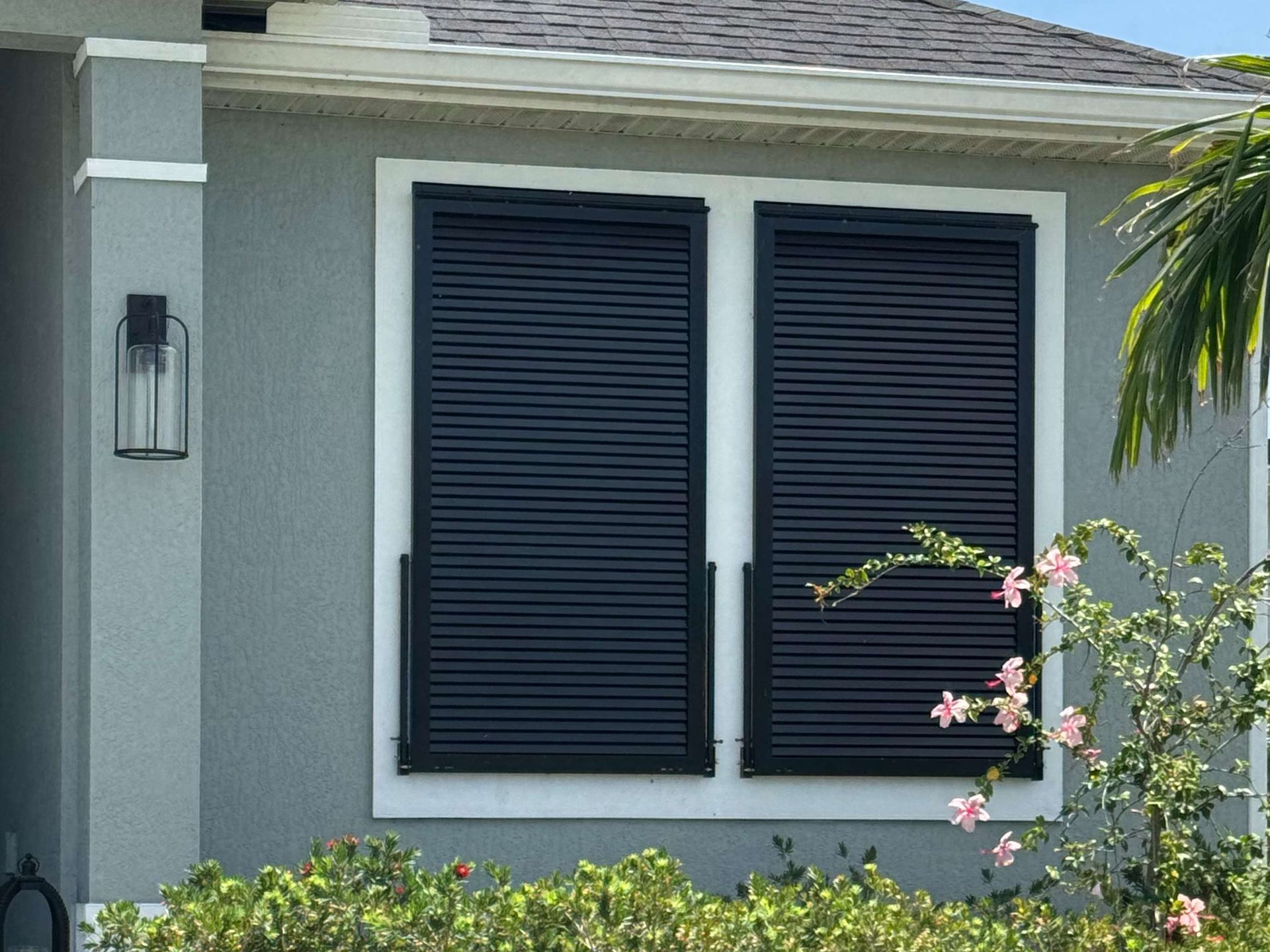 A grey house exterior featuring a window covered by two closed, black horizontal-slat shutters, with a light fixture nearby.