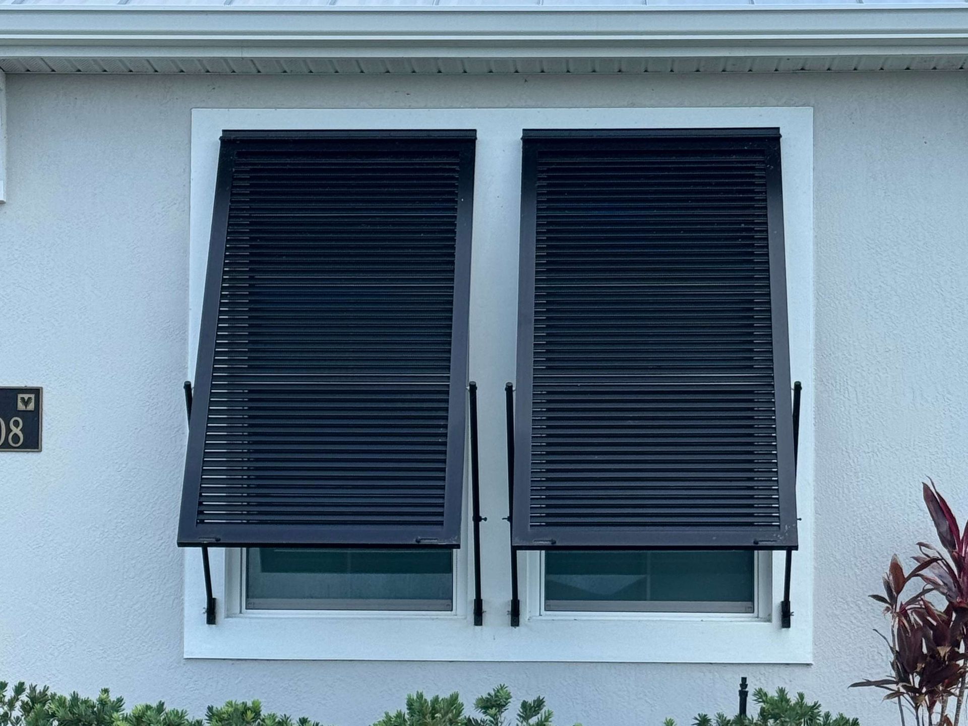Two black louvered storm shutters propped open over a double-pane window on a light-colored stucco wall.