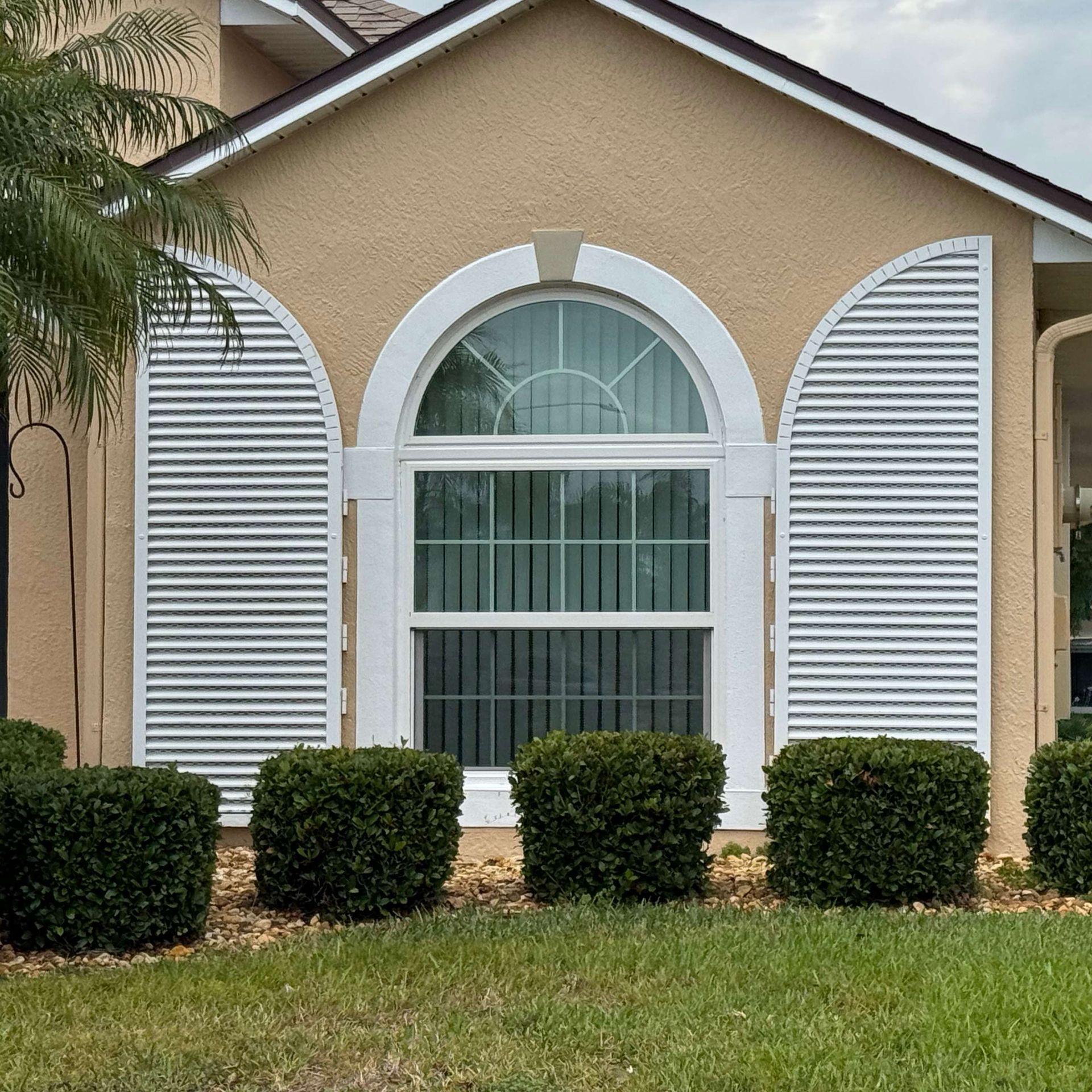 A light tan house exterior features a central arched window with white frames and decorative white shutters on each side.